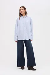 Camisa de corte oversize con diseño de rayas verticales finas en tonos claros. Presenta cuello clásico, cierre frontal con botones y mangas largas con puños abotonados.