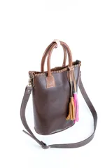 Bolso matera de cuero color marrón con doble asa de mano y correa larga desmontable. Incluye accesorio decorativo de borlas rosas y blancas.