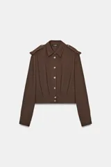 Camisa corta de color marrón con cuello solapa y manga larga. Presenta trabillas en los hombros, pliegues delanteros y cierre frontal con botones.