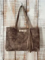 Cartera tipo tote de gamuza en color marrón, con diseño rectangular, doble asa de hombro y cierre frontal metálico giratorio. Incluye un detalle de borla colgante en el lateral.