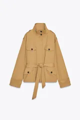 Chaqueta trench corta color beige, con cuello subido, manga larga con trabillas en hombros y mangas, solapas en pecho, bolsillos con solapa en delantero, cinturón lazada en mismo tejido, forro interior y cierre frontal con botones ocultos por solapa.