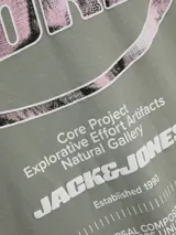 Remera verde militar de algodón con cuello redondo y manga corta. Estampado en la espalda con diseño ovalado que contiene la palabra 'CORE' y texto con la leyenda 'Core Project Explorative Effort Artifacts Natural Gallery JACK&JONES Established 1990'.