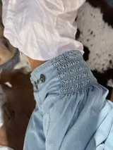 Pantalón de denim celeste claro, con cintura alta fruncida con elástico (smock) y dos botones decorativos en el lateral superior. El tejido es de algodón 100%.