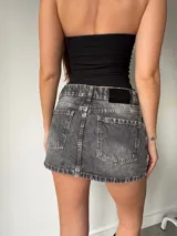 Minifalda de jean rígido color negro con efecto lavado acid wash, tiro bajo y diseño de bolsillos delanteros superpuestos con detalles de tachas metálicas.