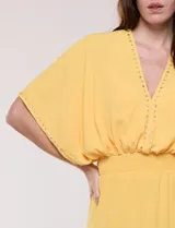 Vestido largo amarillo de corte fluido con escote en V, mangas cortas amplias y apliques de tachas en el escote y puños. Posee cintura elastizada fruncida y falda larga con tajos laterales en el ruedo.