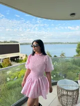 Vestido corto de tela cuadrillé en tono rosado, con cuerpo fruncido tipo nido de abeja, cuello alto y mangas cortas abullonadas. Presenta una falda con vuelo y corte evasé.