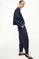 Pantalón de tiro alto confeccionado en denim oscuro, con pinzas delanteras que aportan volumen y pernera abullonada. Cuenta con bolsillos frontales, falsos bolsillos traseros con solapa y bajo ajustable mediante botones a presión.