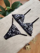 Conjunto de lencería compuesto por un corpiño triangular y una bombacha tipo tanga, confeccionados en encaje negro con detalles florales en hilo plateado.