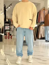 Sweater de punto liviano color beige, con textura marcada, cuello redondo y mangas largas de corte relajado, con pequeño estampado tipográfico negro en el lateral inferior.
