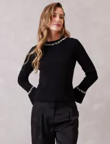 Buzo de tejido soft color negro, con cuello a la base y mangas largas. Presenta apliques de perlas decorativas en el contorno del cuello y en los puños.