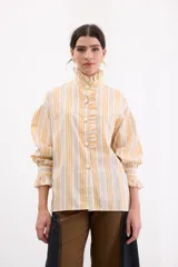 Camisa de corte holgado con estampado de rayas verticales en tonos amarillos, naranjas y beige. Presenta cuello alto con volados, cierre frontal con botones dorados texturados y mangas largas abullonadas con puños terminados en volados.