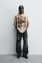 Camiseta de algodón color beige, corte relaxed fit, con cuello redondo y manga corta. Presenta un estampado gráfico grande en negro con motivos variados como una estación de servicio antigua, un caballo alado, una polilla, un pájaro y el texto "RUSTED ROSE".