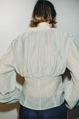 Camisa de manga larga con cuello redondo y escote en pico. Presenta un diseño a rayas verticales en tonos marrones y celestes, con detalle de pliegues en el delantero y los hombros. Cierre frontal con botones y puños ajustados.