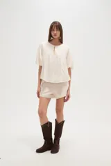 Blusa blanca de manga corta con escote redondo y abertura frontal con botón. Presenta bordados florales en tono beige distribuidos por toda la prenda.