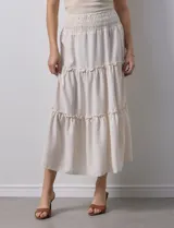 Falda larga de poliéster color beige con finas rayas verticales. Presenta una pretina ancha elastizada con efecto smock y volados en los ruedos de los tres paños que componen la falda.