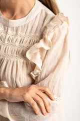 Blusa de color arena con diseño de volados en los hombros, detalle de fruncido en el pecho y mangas largas con puños elásticos.