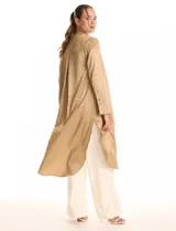 Túnica satinada color dorado con estampado animal print, de silueta oversize y fluida, largo maxi. Cuello a la base con abertura central y botones a tono. Manga larga de corte recto con puños amplios y cierre de botones. Espalda con canesú horizontal recto del cual nace un tablón central profundo. Bajo asimétrico redondeado con tajos laterales profundos, el largo de espalda mayor al largo delantero.