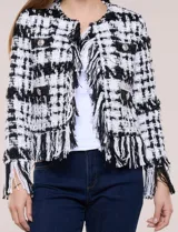 Chaqueta de tweed en blanco y negro con patrón de cuadros, escote redondo, bolsillos frontales con botones metálicos y flecos decorativos en los bordes y puños.