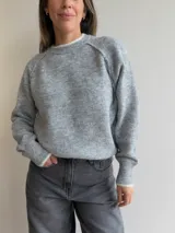 Sweater de punto de corte holgado en color gris melange, con cuello redondo y puños acanalados que presentan un detalle de contraste en color blanco. Posee mangas tipo raglán y una textura suave y elástica.