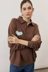 Buzo marrón de manga larga, cuello polo con botones y detalle de aplicación verde en el pecho.