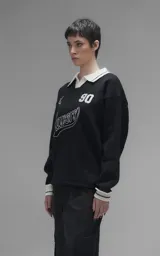 Sweater de punto negro con cuello polo en color crema, manga larga y diseño oversize. Presenta bordados en el frente con la inscripción 'Ravers', el número '90' y letras cursivas, con terminaciones en punto rib en puños y cuello.