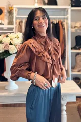 Blusa de color marrón con diseño romántico, cuello alto con lazo ajustable y volados tipo shabot en el frente. Presenta mangas largas con puños fruncidos y una textura liviana.