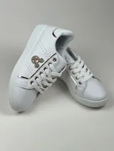 Zapatillas deportivas de cuero sintético en color blanco, beige y negro, con cordones y aplique decorativo dorado con forma de moño.
