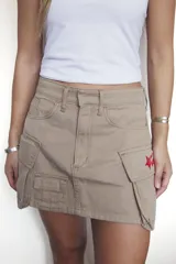 Falda corta estilo cargo color beige, con bolsillos laterales y un parche bordado rojo con una estrella y texto en el bolsillo derecho.