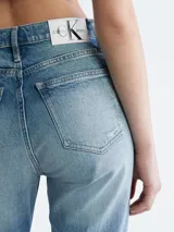 Jean de mezclilla azul claro con efecto lavado y zonas desgastadas/rotas en ambas piernas. Presenta un corte mom de talle alto, estilo clásico de cinco bolsillos y cierre de botón y cremallera.