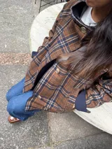 Campera reversible de corte amplio y cuello alto. Un lado presenta un diseño a cuadros en tonos marrones y azules, mientras que el otro lado es de color azul liso. Cuenta con cierre frontal de botones y mangas largas.