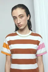 Remera de manga corta con diseño a rayas horizontales en blanco y marrón, con detalles de costuras decorativas en zig-zag de color verde. Las mangas presentan bloques de color naranja en un lado y rosado en el otro.