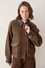Campera corta marrón de tejido texturizado con detalles en cuerina marrón oscura en el cuello, puños, solapas de los bolsillos y ribetes. Cuenta con bolsillos frontales con solapa y botones dorados metálicos, y cierre frontal oculto.