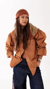 Camisa de color camel con cuello clásico, cierre frontal de botones de madera y mangas largas abullonadas con puños ajustados. Presenta una silueta relajada y versátil.