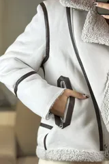 Campera tipo gamulán de color crema con interior y cuello de corderito sintético. Presenta cierre frontal cruzado, bolsillos laterales con cierre y detalles de costuras en contraste color marrón oscuro.