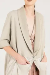 Blazer de lino color beige, de corte holgado y estilo kimono, con mangas cortas caídas, cuello chal, un botón de cierre y bolsillos frontales.
