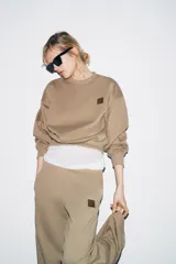 Sudadera de algodón color beige, con cuello redondo, manga larga y corte ligeramente corto. Presenta un pequeño parche cuadrado marrón con un logo grabado en el pecho.