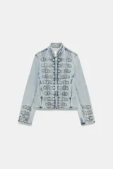 Chaqueta de denim con efecto lavado, corte entallado y cuello redondo. Presenta un diseño de estilo militar con cierre frontal de cremallera metálica y aplicaciones decorativas de botones y costuras en el delantero y los puños.
