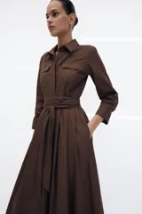 Vestido midi de estilo camisero color verde oscuro, con cuello solapa, escote en pico y cierre frontal con botones. Presenta manga larga, bolsillos laterales ocultos, solapas decorativas en el pecho y cinturón de lazada en el mismo tejido.