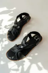 Sandalias chatas de cuero negro con diseño de tiras cruzadas en el empeine y ajuste elástico en el talón. Presentan una plantilla acolchada y suela plana de color natural.