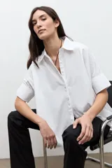 Camisa blanca de corte oversized, confeccionada en algodón, con cuello clásico, cierre frontal de botones y mangas cortas con vuelta.