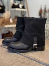 Bota corta de diseño combinado en color negro, con puntera y base de cuero liso y caña de gamuza con efecto arrugado. Cuenta con una tira lateral con hebilla metálica plateada.