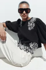 Blusa corta negra con mangas abullonadas y bordados florales en contraste de color blanco. Presenta cuello redondo con puntilla, mangas ajustables con cordones y borlas, y cierre en la espalda con abertura y botón oculto.