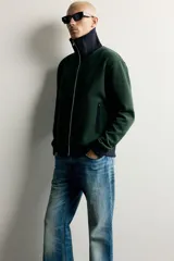 Chaqueta regular fit color verde oscuro con detalles en negro en el cuello subido, los puños, el dobladillo inferior y los ribetes de los bolsillos. Cuenta con cierre frontal de cremallera metálica y bolsillos con cierre en la cadera.