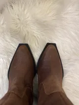 Bota estilo texana de caña media, confeccionada en gamuza color marrón. Presenta puntera ligeramente cuadrada, taco cuadrado de altura media en color negro y costuras reforzadas en el diseño de la caña.