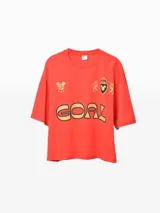 Remera naranja con cuello polo blanco, estampado frontal con la palabra "GOAL" en amarillo y negro, y logos bordados.