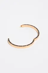 Pulsera rígida de metal dorado con detalles de esmalte negro en el borde y letras grabadas a lo largo de la pieza. Cuenta con cierre de bisagra.