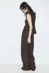 Pantalón sastrero de tiro alto, color marrón oscuro, con pinzas frontales y raya marcada en las piernas. Presenta un corte amplio y recto.