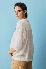 Camisa blanca de lino, con mangas anchas a tres cuartos, cuello clásico, cierre frontal con botones y detalles de calados verticales en la parte central y en los puños.