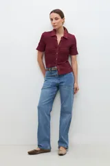 Camisa de manga corta en color bordó, confeccionada en tejido con textura de punto smock. Presenta cuello clásico, cierre frontal con botones y un ajuste entallado al cuerpo.