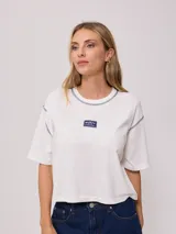 Remera blanca de algodón con corte cropped y calce holgado. Tiene cuello redondo con ribete azul y logo de la marca estampado en el pecho.